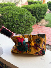 Load image into Gallery viewer, A multicolor floral velvet makeup pouch with a yellow base, resting on a white ledge with a pink hairbrush resting inside it. The background shows green hedges and a brick pathway.