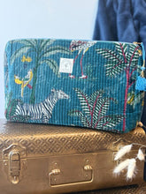 Load image into Gallery viewer, A rectangular quilted toiletry bag with a blue jungle animal print sits on top of a vintage gold suitcase. The bag features a zebra, flamingos, and palm trees in teal, green, pink, and yellow, with a small white brand label on the front.
