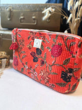 Load image into Gallery viewer, A red quilted fabric cosmetic pouch with a floral tapestry pattern, featuring a top zipper, a red tassel, and a small white brand label, displayed on a white surface with vintage suitcases in the background.
